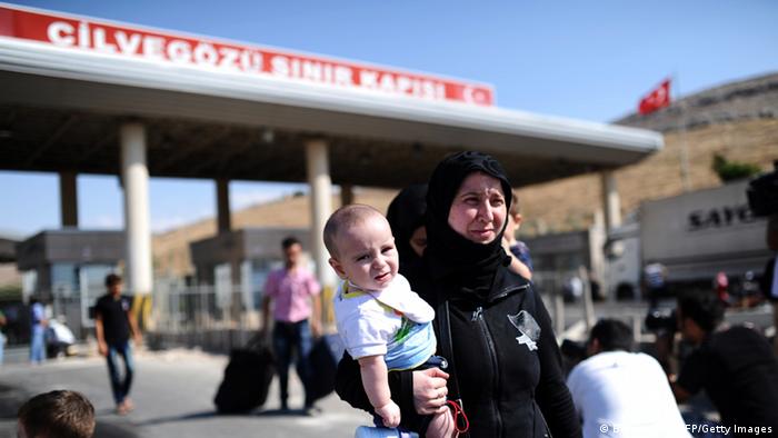 A woman of Middle-Eastern descent wearing a black headscarf and robes carries a small child through an outdoor customs gate (Photo: Bulent Kilic)