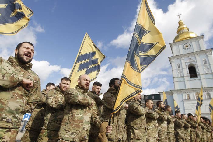 Men in military garb stand with flags featuring the Azov movement's symbol, which is similar to the Wolfsangel, widely used by Nazi German divisions during the Second World War