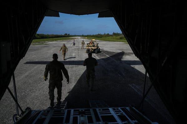 A view from inside a cargo plane’s open hatch looking down a runway where some soldiers and a military vehicle are visible.