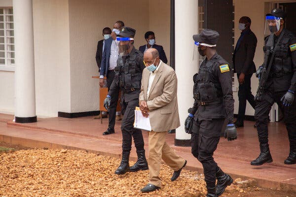 Paul Rusesabagina, wearing a tan jacket, white shirt and tan trousers, escorted by uniformed police officers out of a white building with columns.
