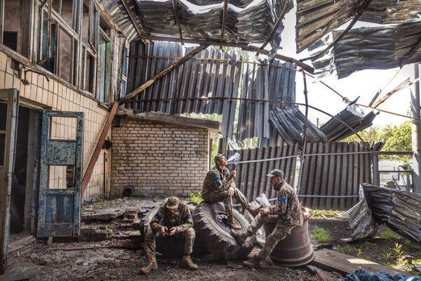 Three men in camouflage, with blue armbands, sit on large wheels and tires, one smoking and one looking at his phone.