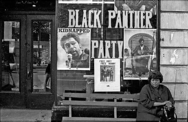 A black-and-white photograph of a woman sitting on a bench outside an office. A large glass window is covered with posters and has “Black Panther Party” painted on it.
