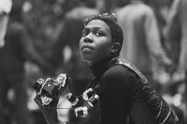 A black-and-white photograph of Afeni Shakur, dressed in a black turtleneck and vest and holding a camera.