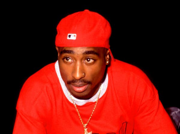 A color photograph of Tupac Shakur, wearing a red ball cap and red shirt.