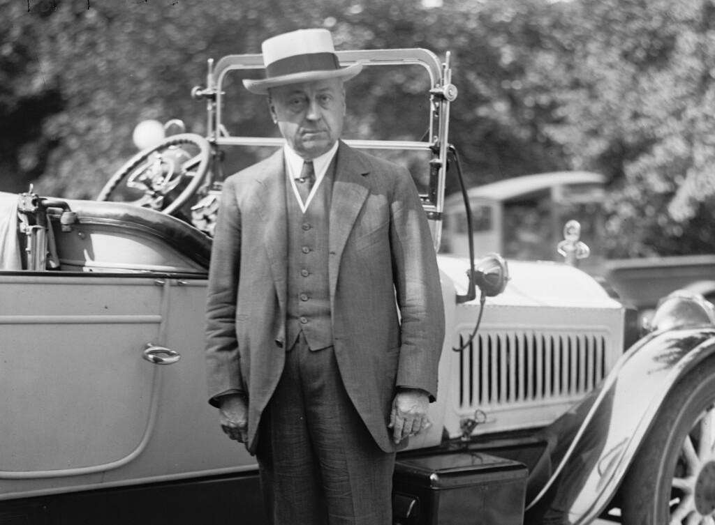 A person in a suit and hat standing next to a car

Description automatically generated with medium confidence