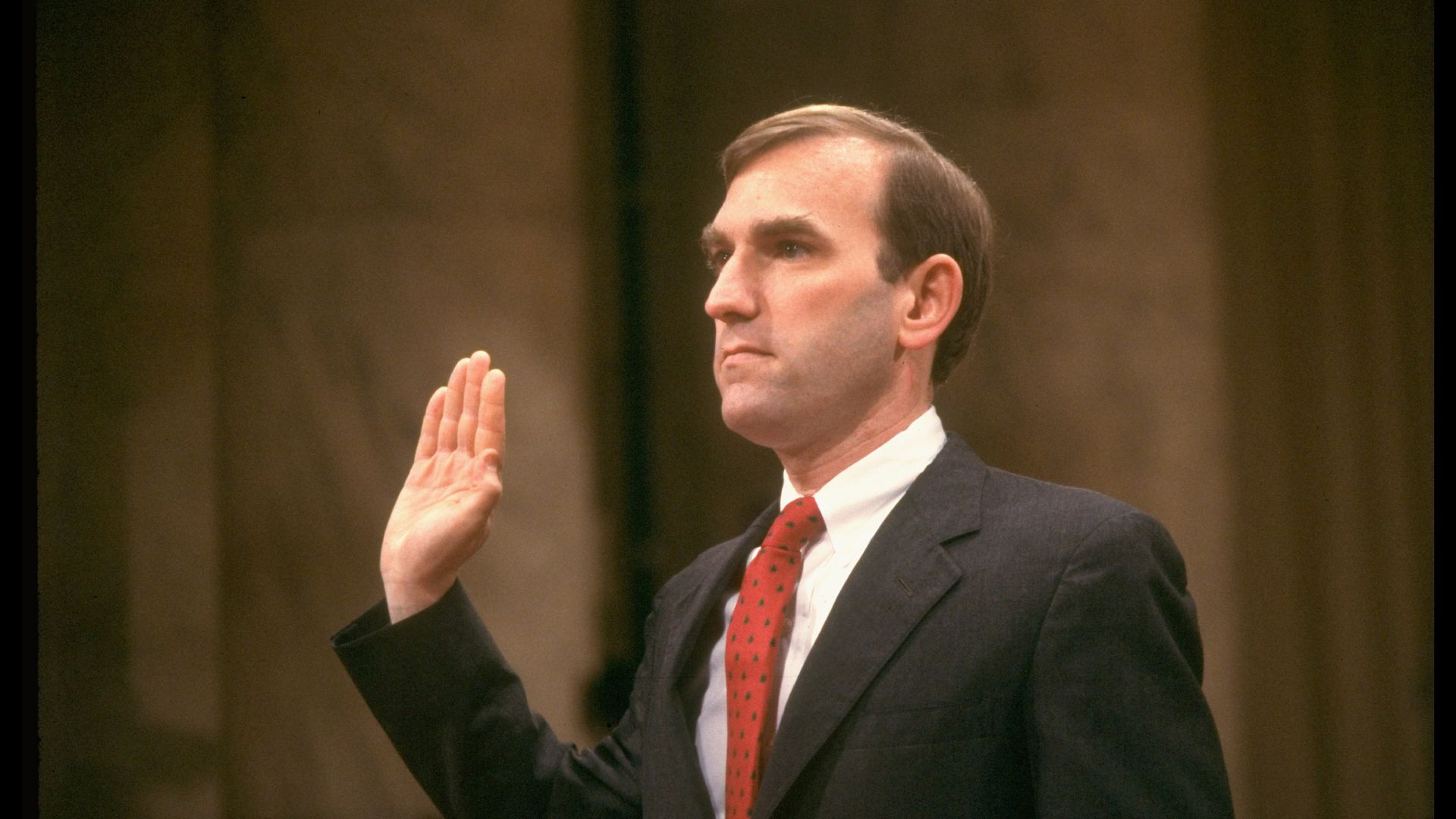 A person in a suit and tie raising his hand

Description automatically generated