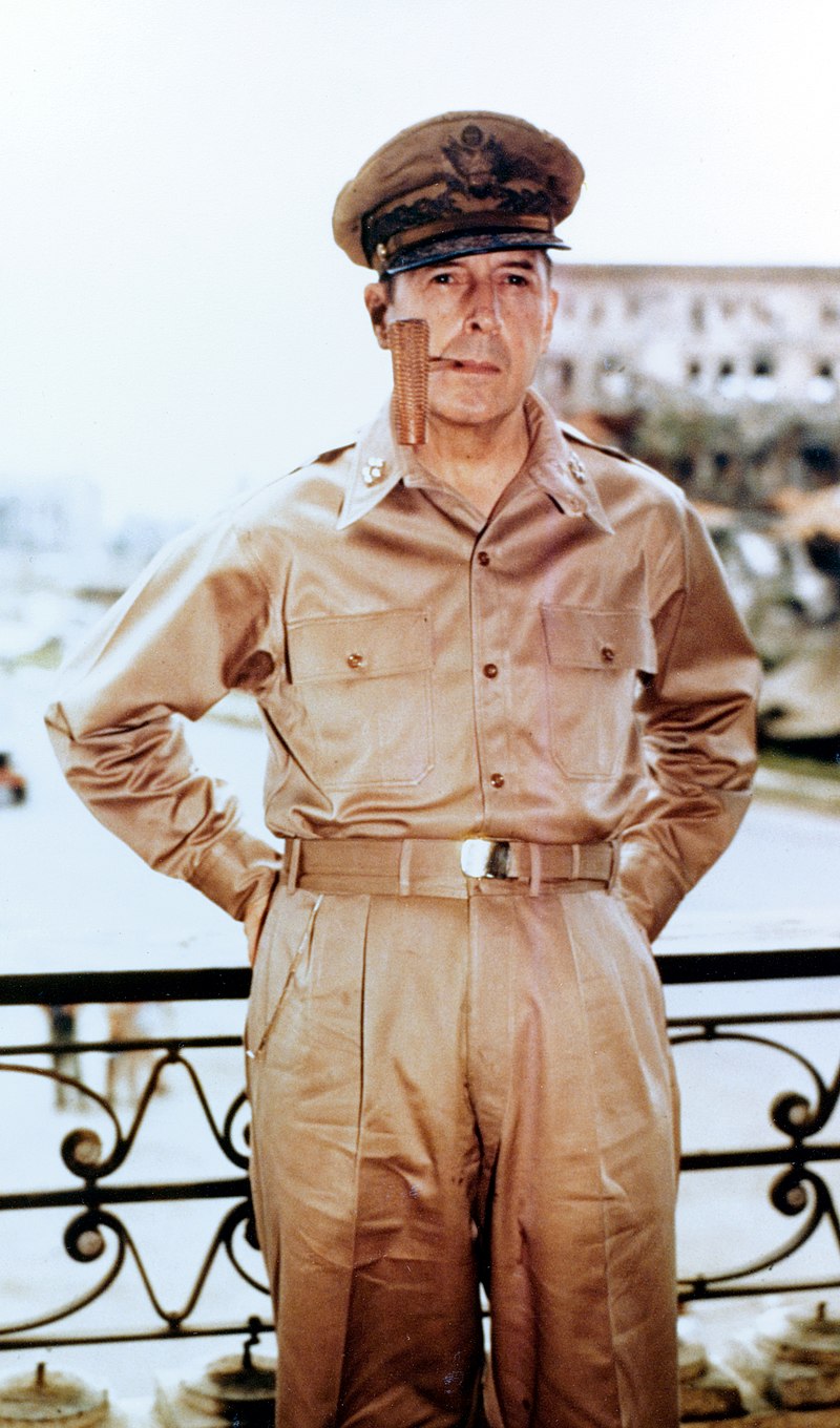 MacArthur in khaki trousers and open necked shirt with five-star-rank badges on the collar. He is wearing his field marshal's cap and smoking a corncob pipe.