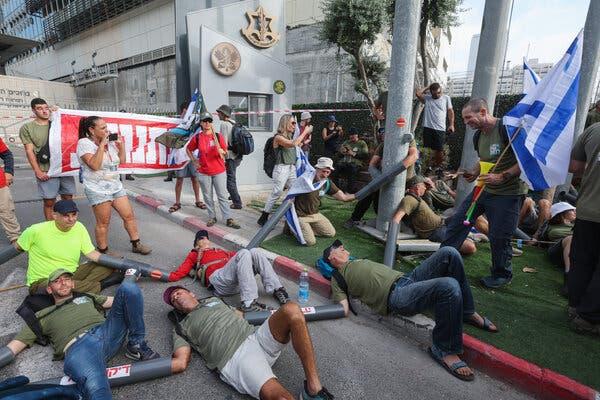 A group of people, some with drainpipes on their arms and over their hands gathered outside an gray building.