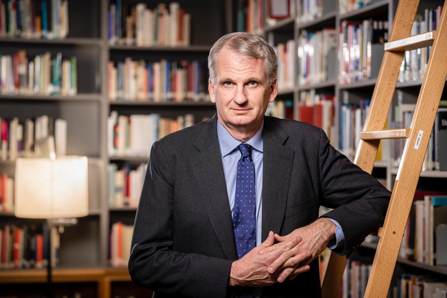 A person in a suit standing in front of a bookshelf

Description automatically generated