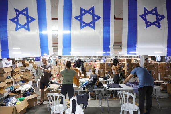 Three Israeli flags hang over people folding clothes in a hall full of cardboard boxes.