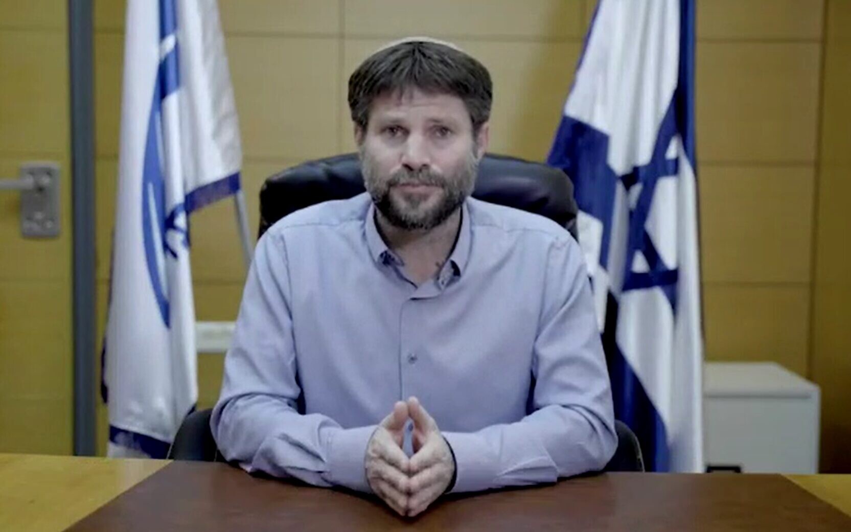 A person sitting at a desk with flags behind him
Description automatically generated