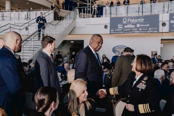 Defense Secretary Lloyd J. Austin III shaking hands with a woman in a military uniform as other people are seated at tables around them.