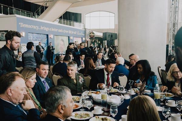 People sitting around a round table eating a meal in a crowded room. Some people are wearing military uniforms and others are wearing suits.