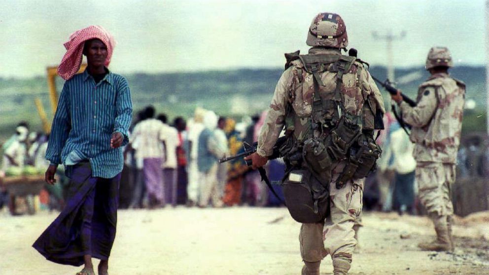 A soldier walking with a person in the background

Description automatically generated