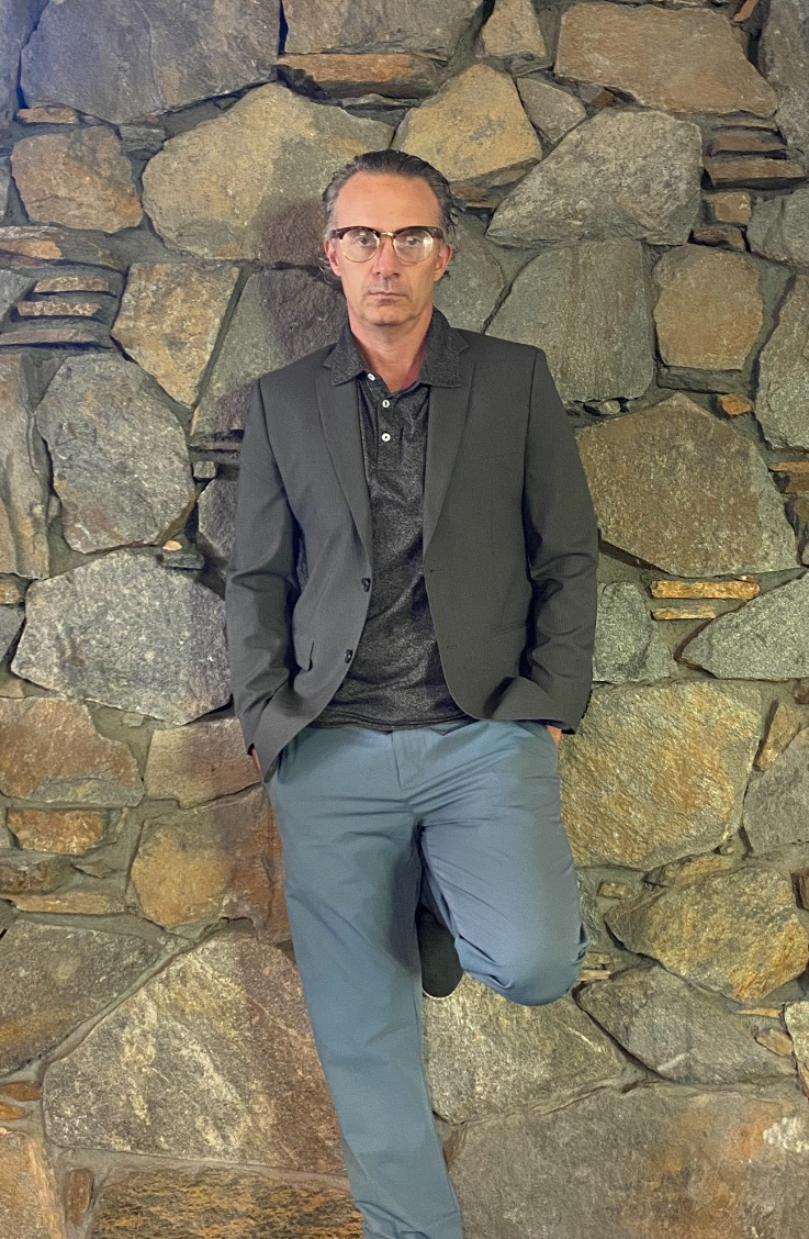 A person in a suit leaning against a stone wall

Description automatically generated