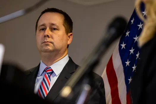A person in a suit standing in front of a flag Description automatically generated