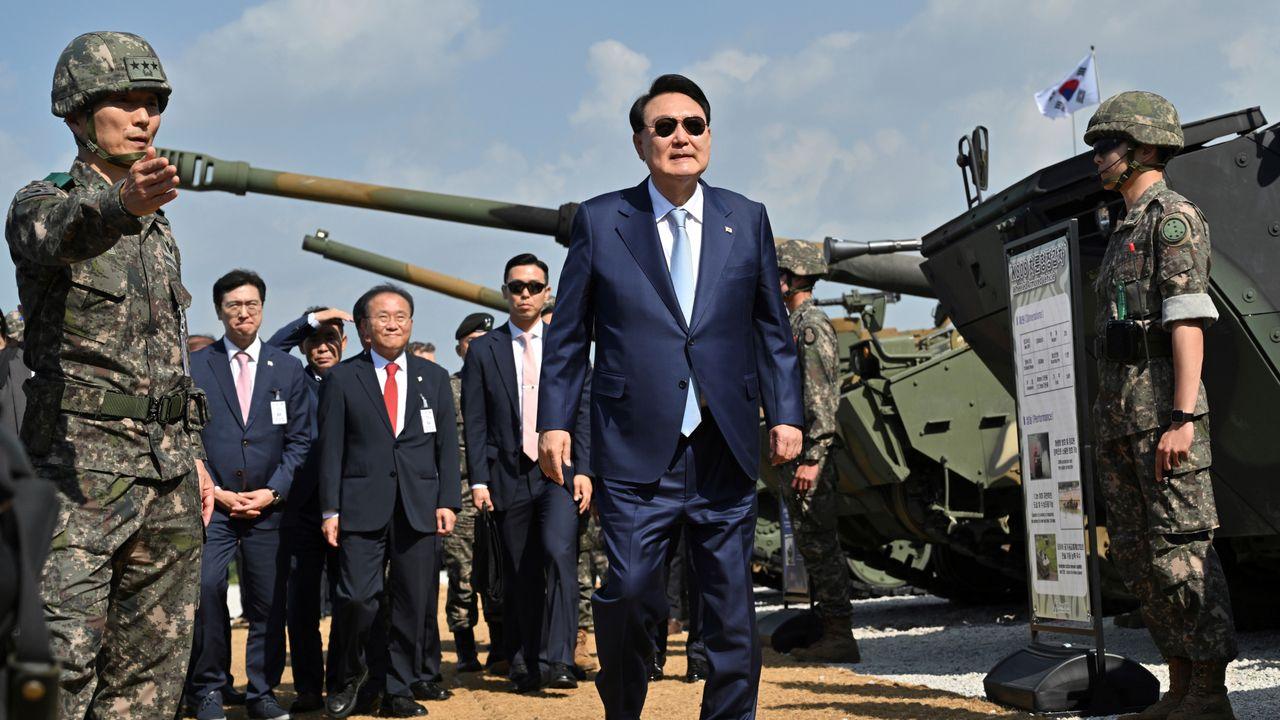 A person in a suit walking in front of military vehicles
Description automatically generated