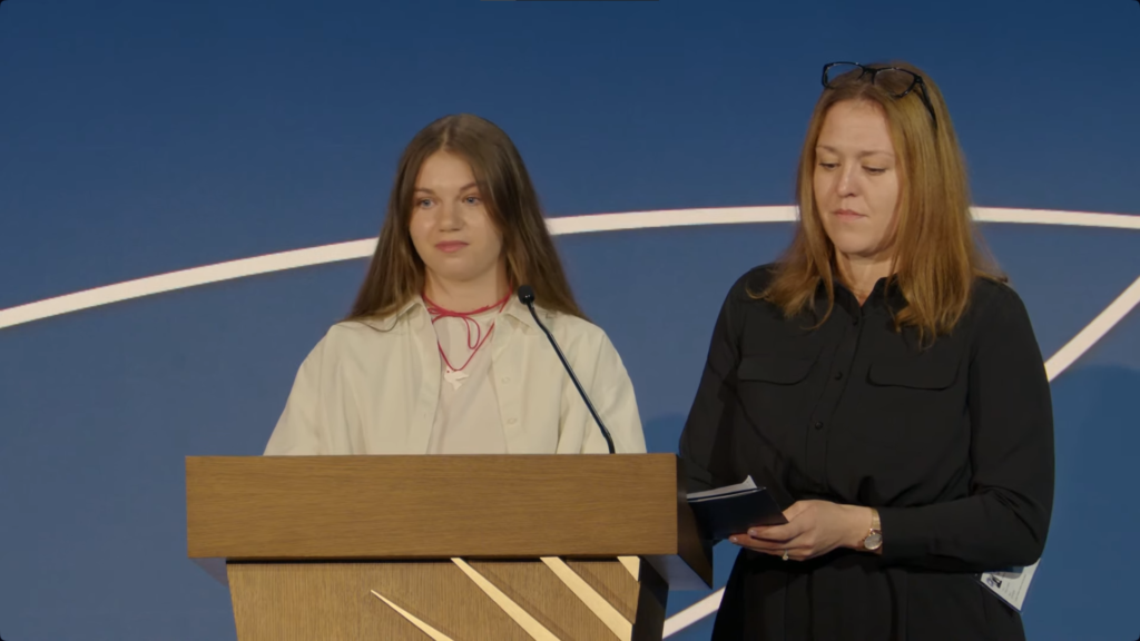 A person standing at a podium with a person standing behind her

Description automatically generated