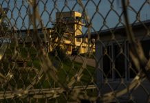 Defense Secretary’s Abrogation of Plea Deal For 9/11 Suspects Will Extend Life of Guantanamo Bay Prison A building with a tower is seen through a chain-link fence.