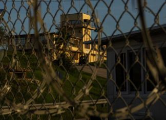 Defense Secretary’s Abrogation of Plea Deal For 9/11 Suspects Will Extend Life of Guantanamo Bay Prison A building with a tower is seen through a chain-link fence.