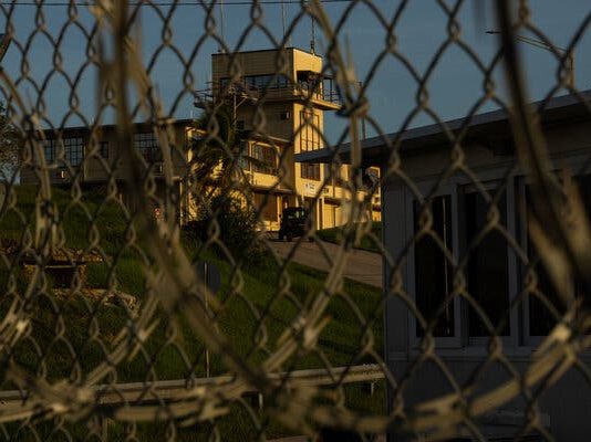 Defense Secretary’s Abrogation of Plea Deal For 9/11 Suspects Will Extend Life of Guantanamo Bay Prison A building with a tower is seen through a chain-link fence.