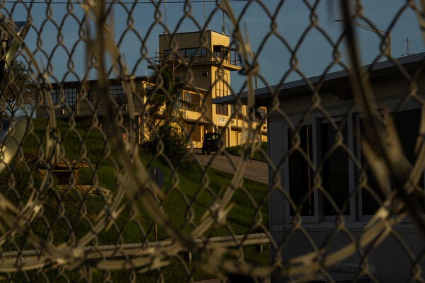 A building with a tower is seen through a chain-link fence.