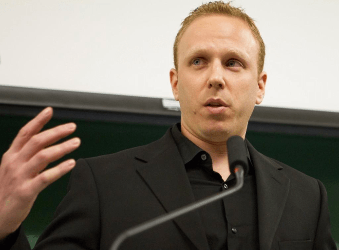 A person in a black suit speaking into a microphone Description automatically generated