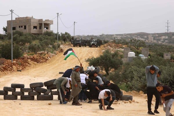 A small group of men stands behind a barricade made of tires, with Israeli military vehicles in the background. One of the men holds a Palestinian flag.