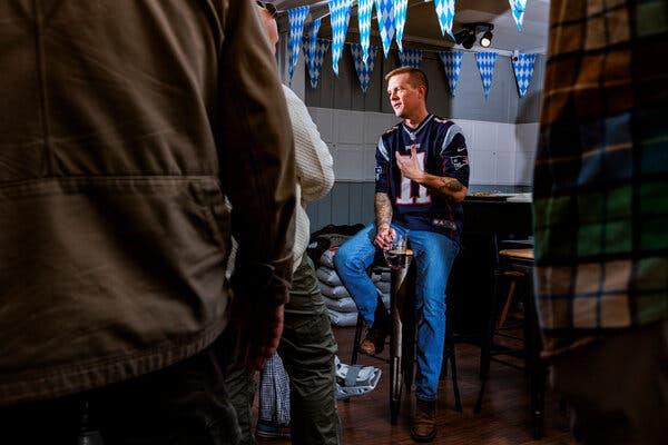 A man in a Patriots football jersey and bluejeans sits on a stool talking and holding a stein of beer.