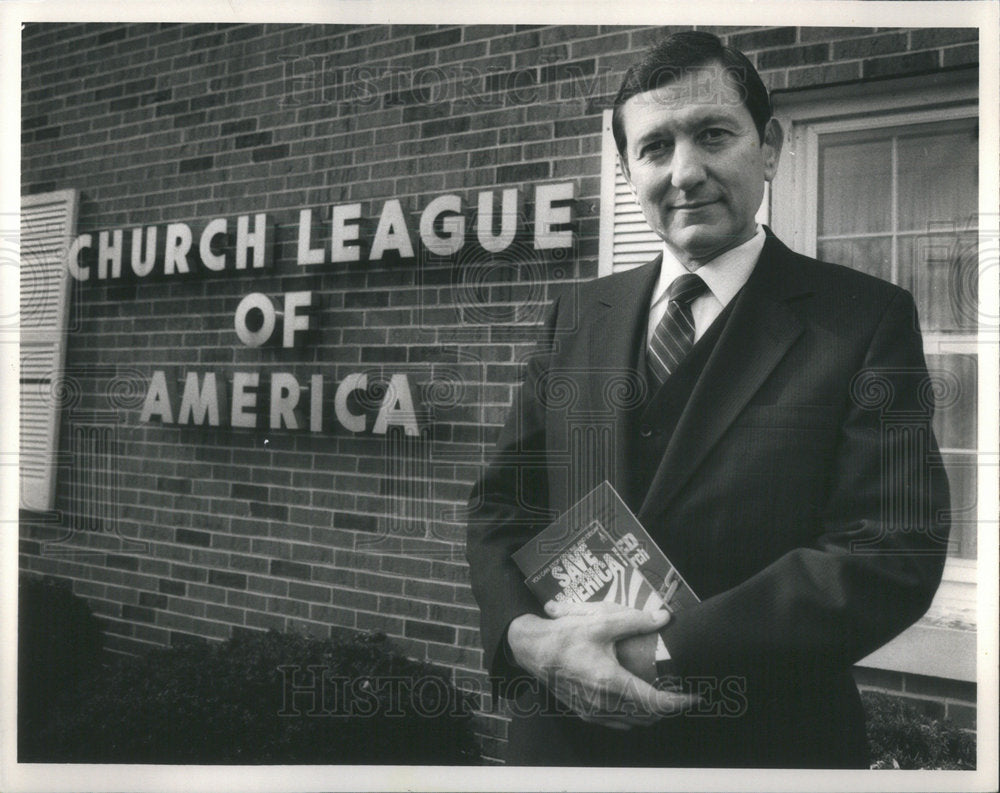 A person in a suit holding a book
Description automatically generated