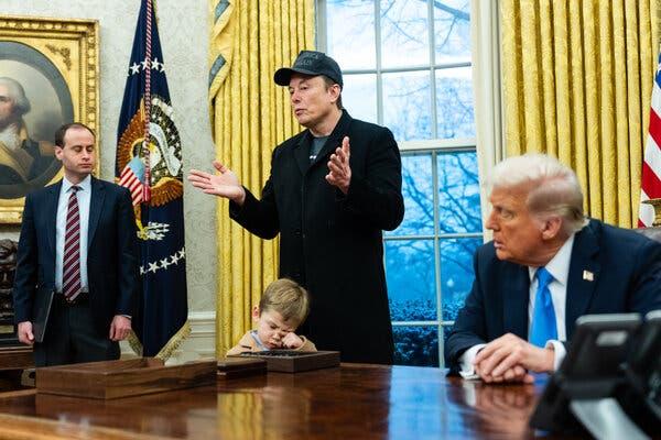 Elon Musk stands against a backdrop of a yellow-curtained window and a portrait of George Washington, holding his hands up, as Donald Trump, seated at a large desk, looks at him.