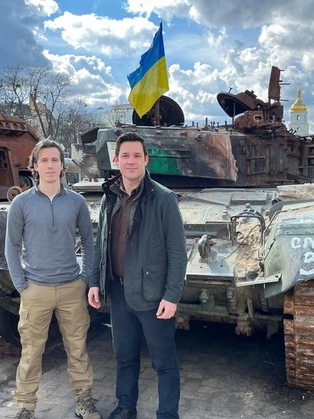 Ian Miller and Evan Platt stand in front of a battered tank. A Ukranian flag flies from the tank.