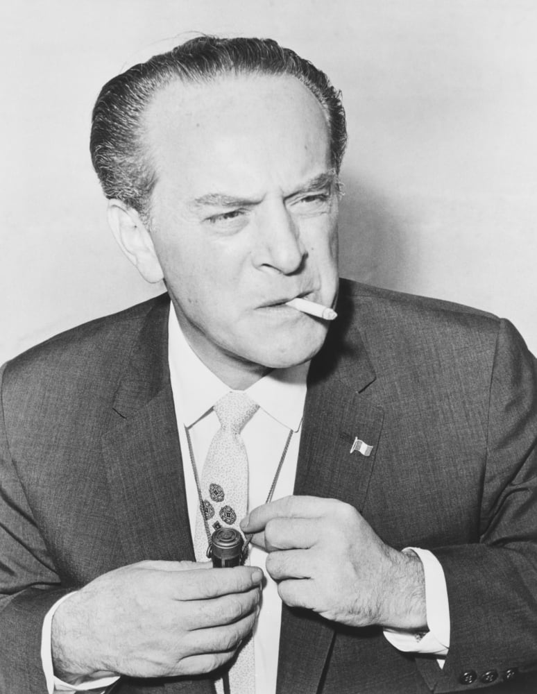 A person in a suit smoking a cigarette
Description automatically generated