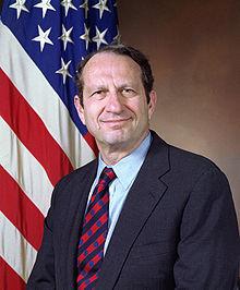 A person in a suit and tie standing in front of a flag

Description automatically generated