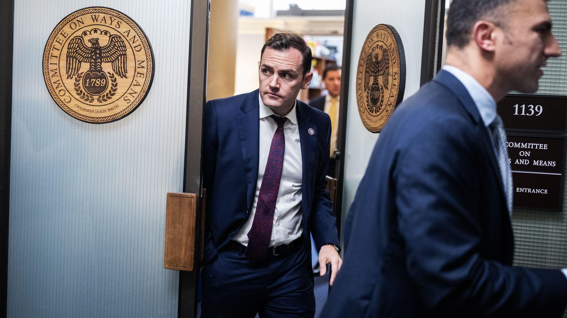 Mike Gallagher, a man in a blue suit and white shirt, steps into a hallway. Two open doors are adorned with the seal of the ways and means committee.