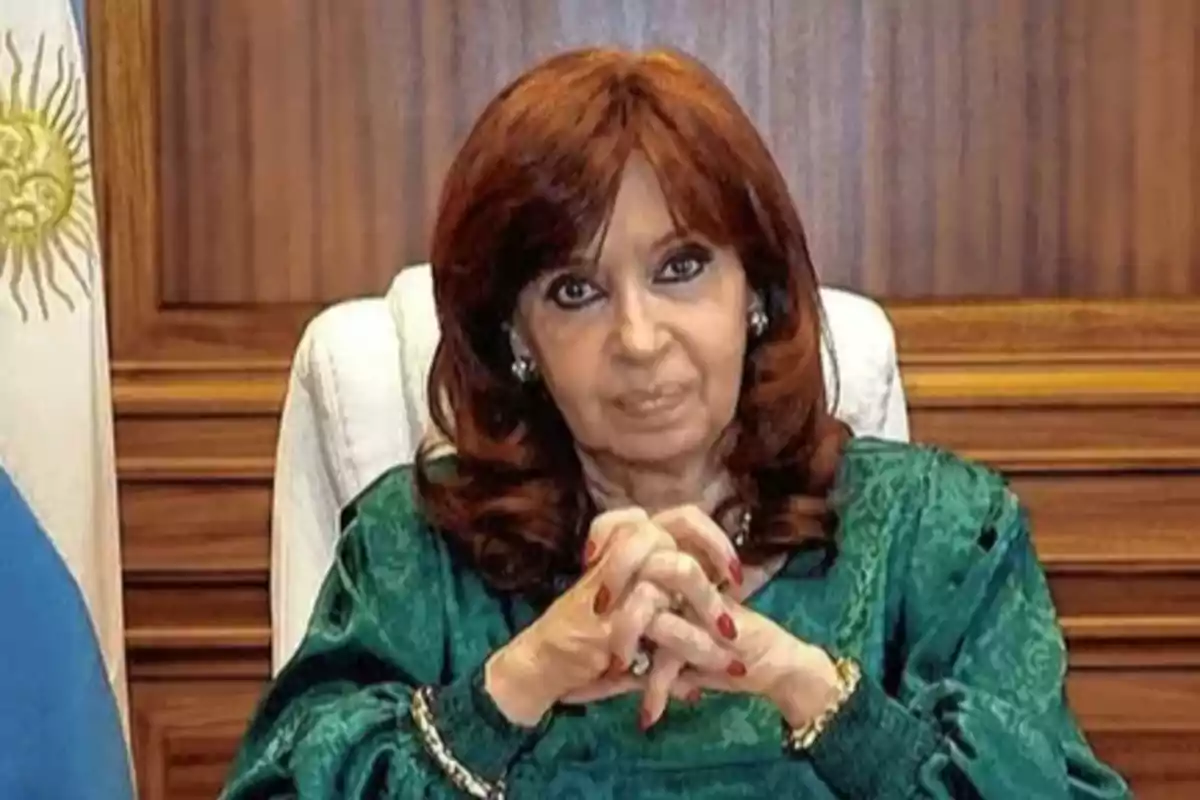 A woman with reddish-brown hair and a green dress is sitting in front of a wooden background with a flag beside her.