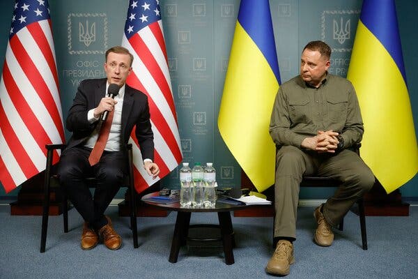 Jake Sullivan, in a black suit and a red tie, speaks into a microphone while seated in front of American and Ukrainian flags. Next to him is Andriy Yermak, the head of the presidential office of Ukraine, who is wearing green military-style clothes.