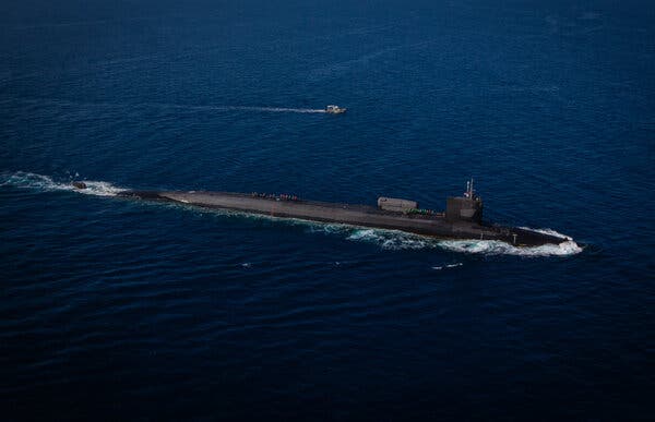 A dark gray submarine crosses open water with a small vessel in the distance. 