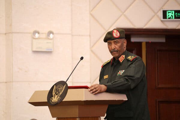 A man in a green military beret and collared shirt with military insignia stands in front of a podium.