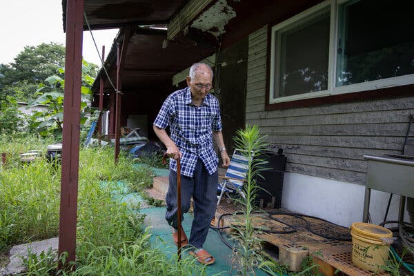 A man with a cane walks along a path outside a wooden building. 