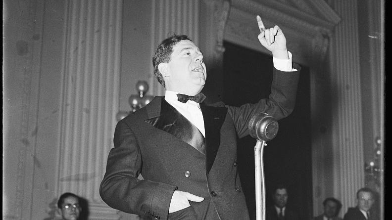 A person in a suit speaking into a microphone
Description automatically generated