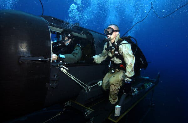 Navy divers in scuba gear are seen underwater, one inside a mini-sub and one next to the mini-sub’s open hatch. 