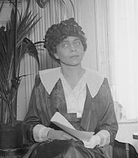 Woman in a dark hat holding a book sits in front of a fronded plant. She is in a white collared shirt, and her head is slightly cocked