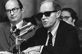 A person in sunglasses sitting at a podium with microphones
AI-generated content may be incorrect.