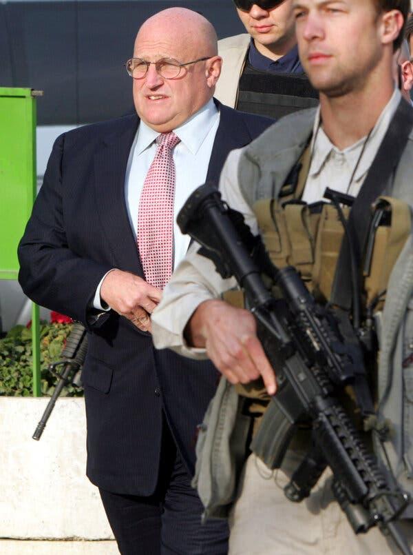 Richard Armitage, a bald man wearing glasses and a suit, stands behind an armed man who is presumably guarding him.