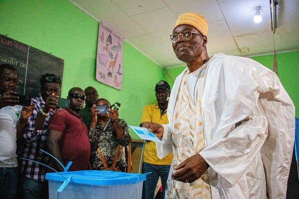 Issa Tchiroma Bakary being photographed while dropping a paper ballot into a bin.