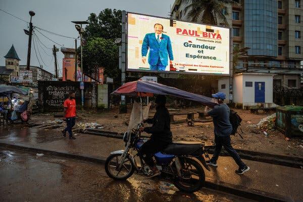 People walking and riding motorcycles in front of an illuminated billboard showing President Paul Biya.