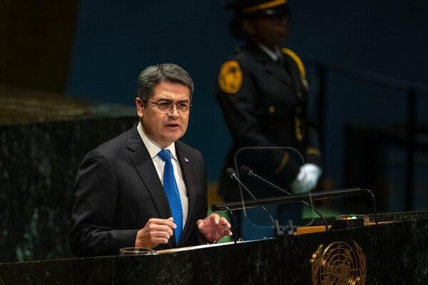 A bespectacled man in a suit and blue tie stands at a lectern. In the shadows behind hm is a uniformed figure with white gloves.