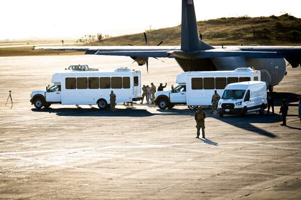 Men in camouflage uniforms lead a man in a gray sweatsuit away from a military plane onto a white bus on a tarmac.