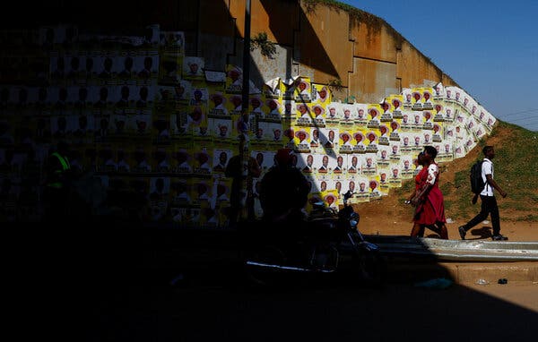 Numerous yellow and white posters line a wall. Three people walk on a path, while a motorcycle and two figures in shadow are in the deep foreground.